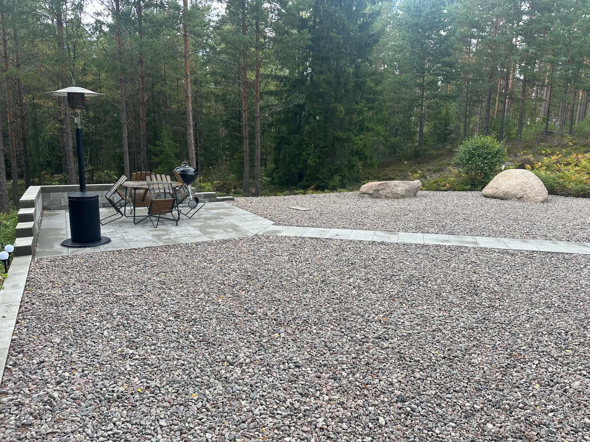 Uteplats med plattor, grusyta och stödmur mot skog